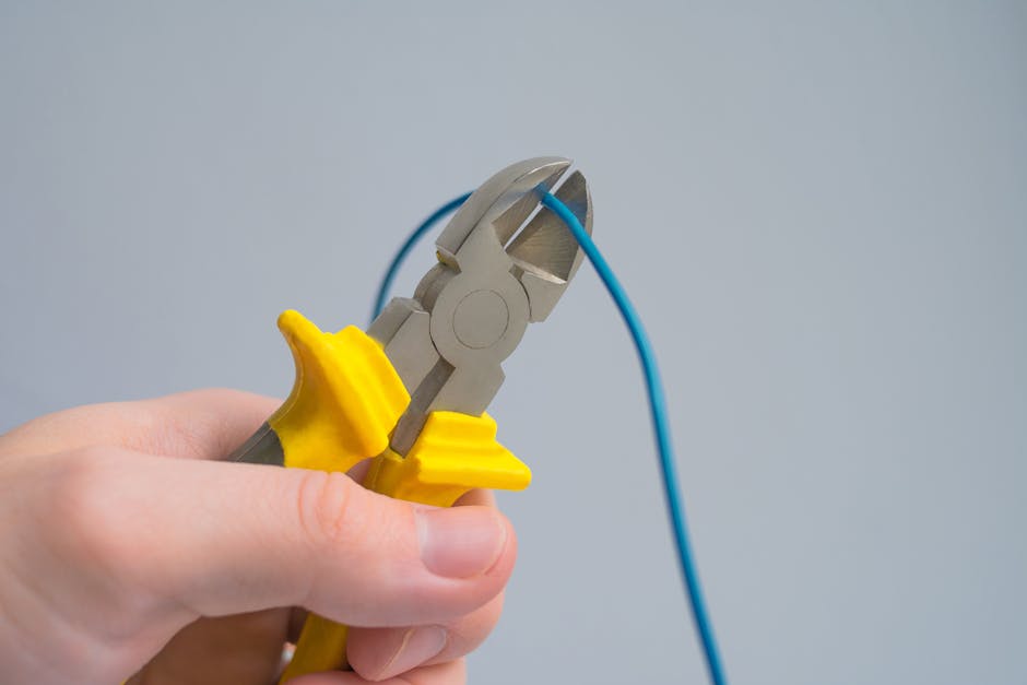 A close-up image of a hand using pliers to cut a blue electrical wire against a gray background.